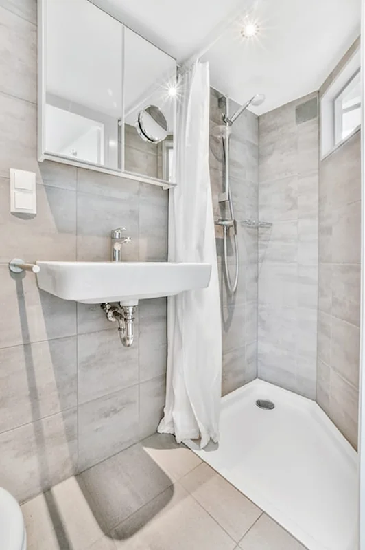 Nowoczesna mała łazienka z szarymi kafelkami, prysznicem w rogu, umywalką, lustrem i lampą